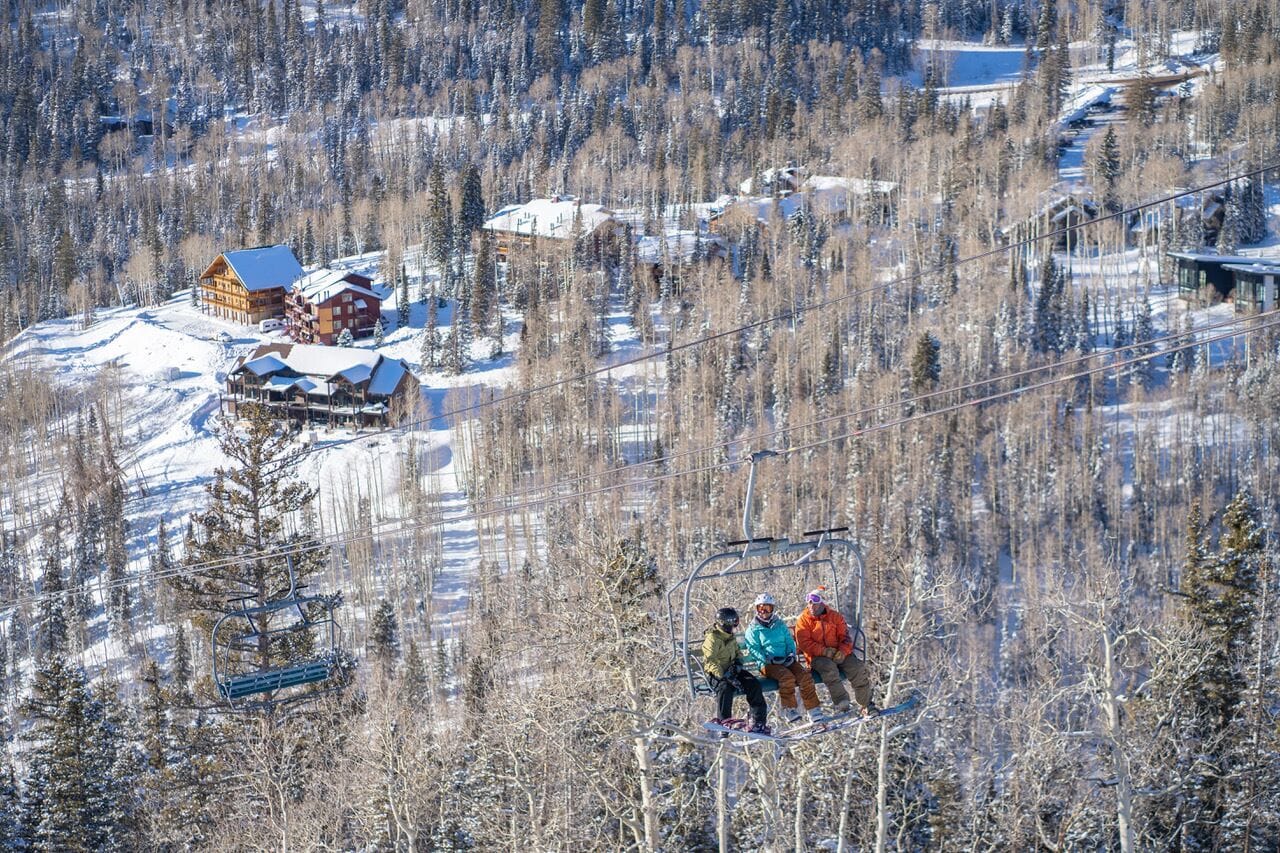 A ski lift with passengers, surrounded by snow-covered trees and buildings.