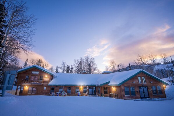 A wooden lodge with a balcony and a snow-covered roof, surrounded by trees.