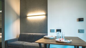 A modern living room with a grey sofa, a wooden dining table with stools, and a wall-mounted light fixture. - Il Piccolo Paradiso - Magnolia - by Be Your Home (Civitavecchia)