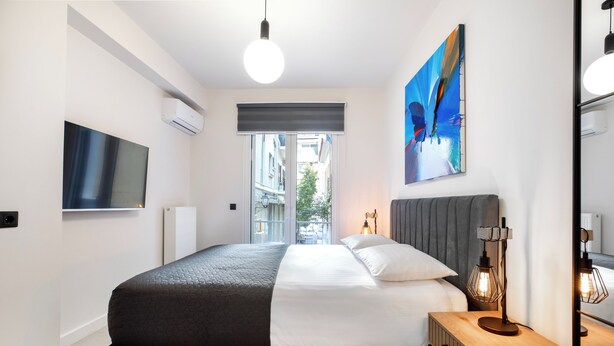 Un dormitorio moderno con una cama grande, una mesita de noche con lámpara, un televisor de pantalla plana y un ventanal con vistas a edificios.