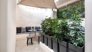 Un pequeño espacio al aire libre con una mesa, sillas y una cubierta. Hay plantas en macetas a lo largo de la pared y un banco con cojines.