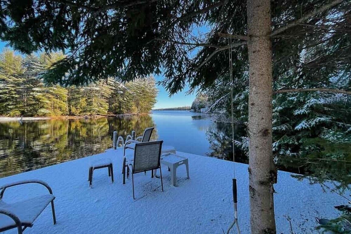 A snow-covered deck with outdoor seating and a view of a lake surrounded by trees.