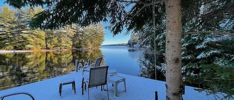 A snow-covered deck with outdoor seating and a view of a lake surrounded by trees.