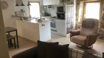 A living room with a sofa, a chair, a kitchen in the background, and a stone wall.