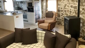 Un salón con sofá, sillón y televisor. Se observa una cocina al fondo, con armarios y electrodomésticos.