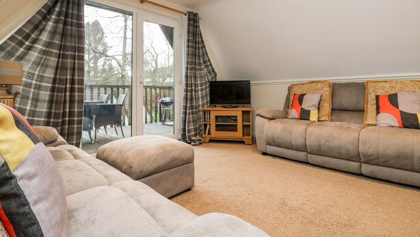 A cozy living room with a sofa, ottoman, and a TV on a wooden stand. A sliding glass door leads to an outdoor deck with a barbecue grill. - Valley Lodge 31 (Gunnislake)