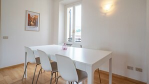 Quarto claro e minimalista com mesa e cadeiras brancas, um quadro na parede e janela com vista.