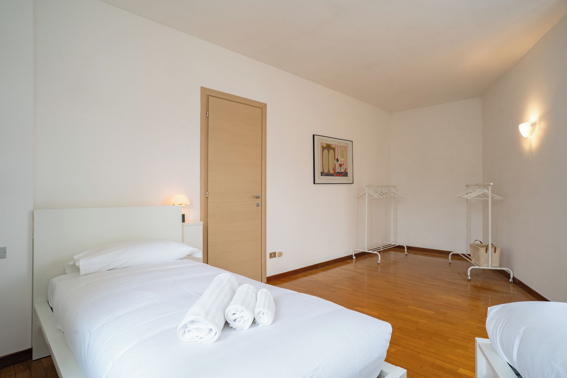 A hotel room with a bed, a wooden door, a wall-mounted picture, and a clothing rack.