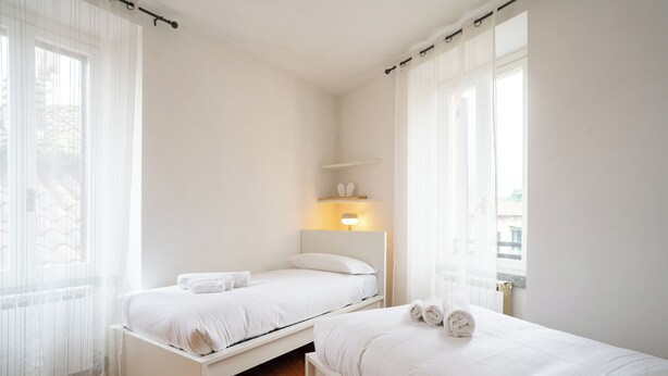 Quarto com duas camas de solteiro, uma mesa de cabeceira com luminária e cortinas translúcidas.