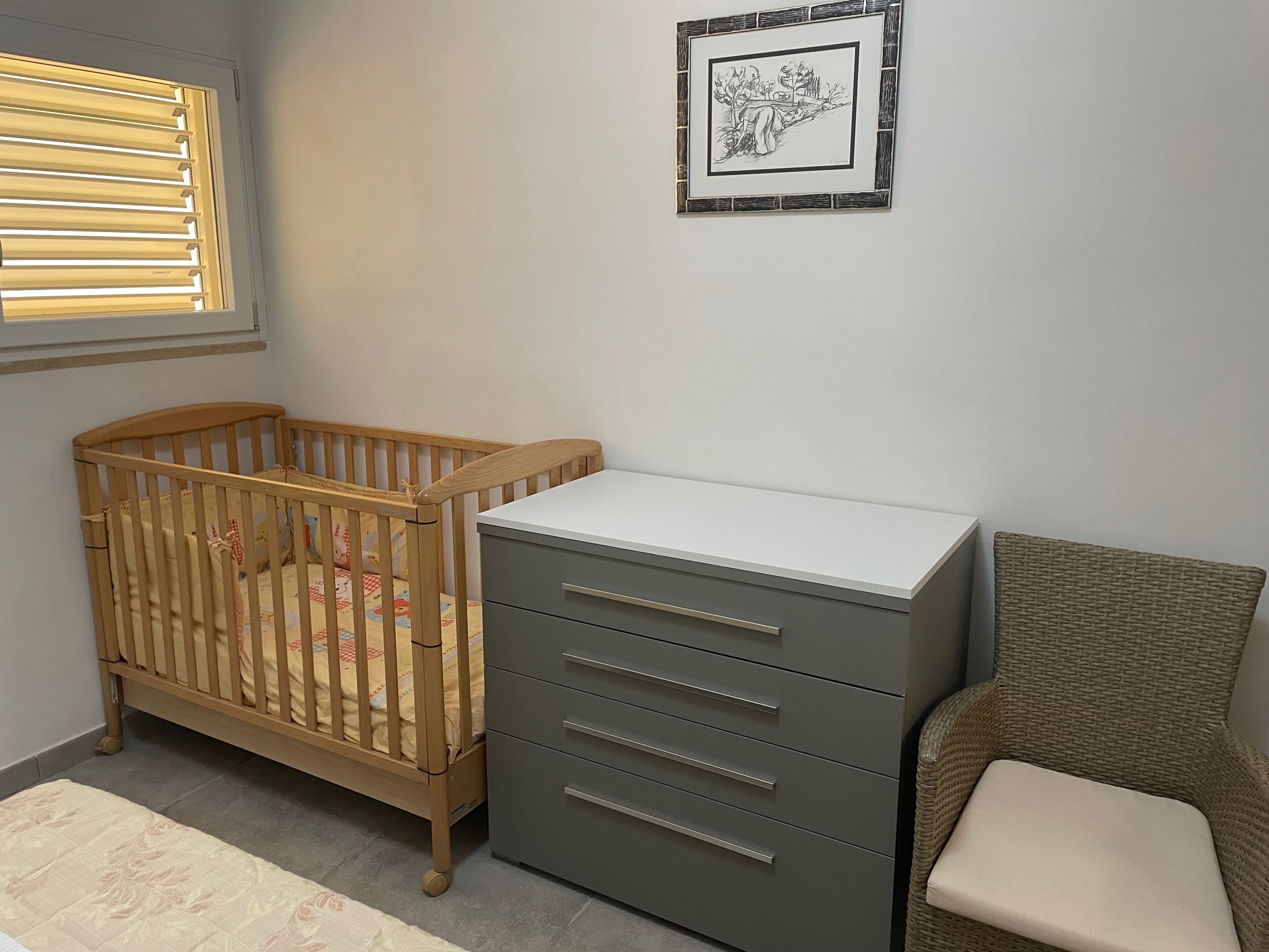 A baby crib, a gray dresser, and a wicker chair in a room with a window and a framed picture on the wall.