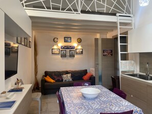Een knus woongedeelte met een bank, een eettafel met een kom, een keukenhoek en een vide met een witte balustrade.