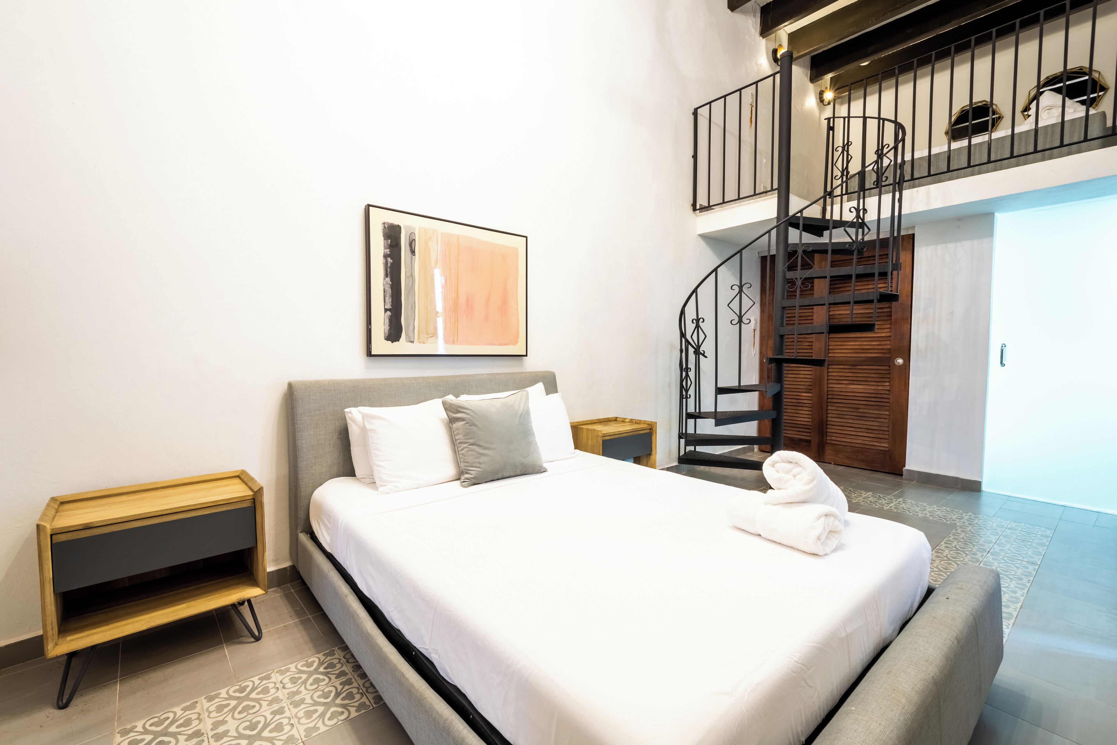 Une chambre moderne avec un grand lit, une table de chevet en bois, un tableau abstrait accroché au mur et un escalier menant à un niveau supérieur.
