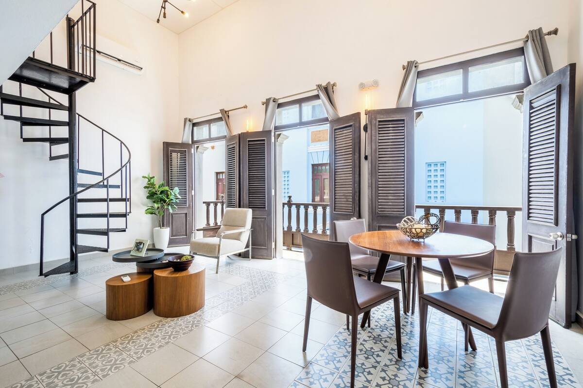 A modern interior with a spiral staircase, a round dining table with chairs, and a view through open shutters to the outside.