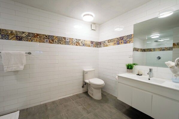 Un baño moderno con azulejos blancos, un inodoro, un lavabo con espejo y un borde decorativo de azulejos.