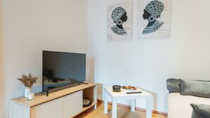 Un salon avec un téléviseur à écran plat posé sur un meuble en bois, une table basse blanche avec une télécommande, et un canapé gris.