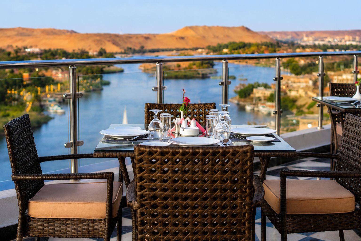 Una mesa de comedor dispuesta con platos, vasos y cubiertos, con vista a un río y colinas.