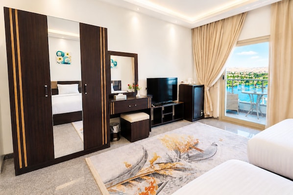 A hotel room with a bed, a TV, a minibar, a desk, a large mirror, and a balcony with a pool view. - Blue Lotus Nile Suites (Aswan)