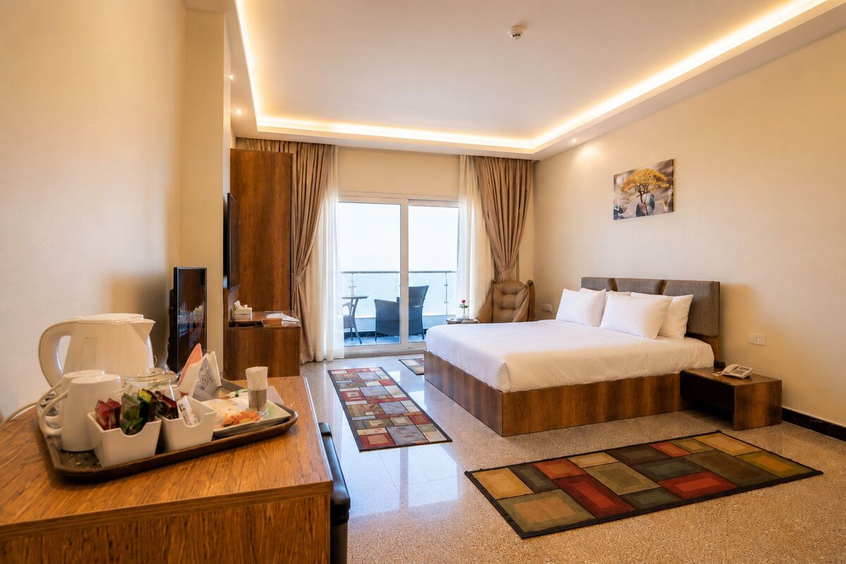 A hotel room with a bed, a desk with a tray of breakfast items, a chair, a small table, and a view of the outside.