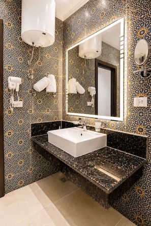 A bathroom with a white sink, a large mirror, towel hooks, and a patterned tile wall. - Blue Lotus Nile Suites (Aswan)