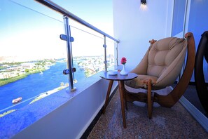 A balcony with a view of a river and cityscape, a cushioned chair, a small table with a vase and a bowl, and a metal railing. - Blue Lotus Nile Suites (Aswan)