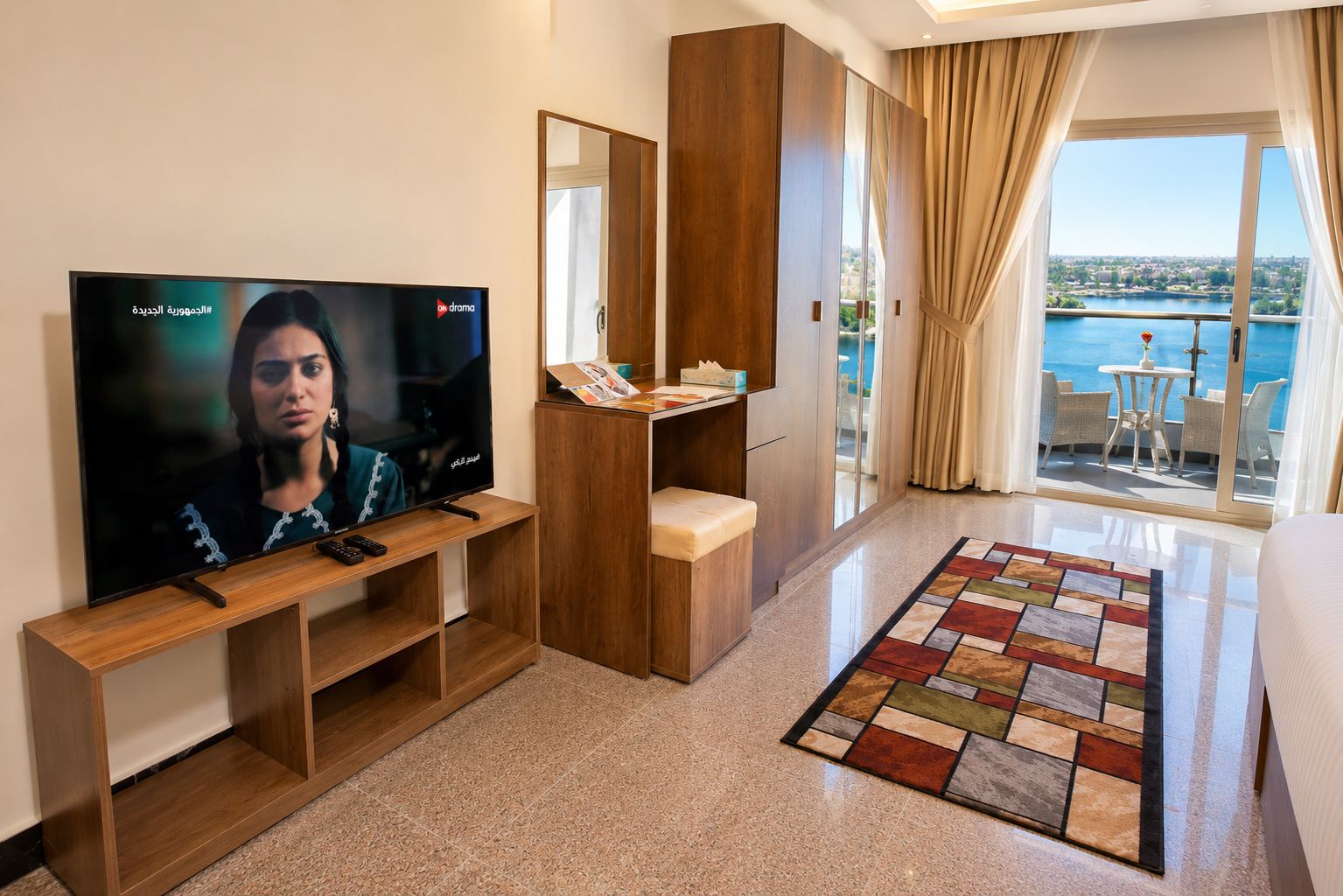 A hotel room with a TV, wooden furniture, a balcony with a view, and a patterned rug.