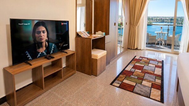 A hotel room with a TV, wooden furniture, a balcony with a view, and a patterned rug.