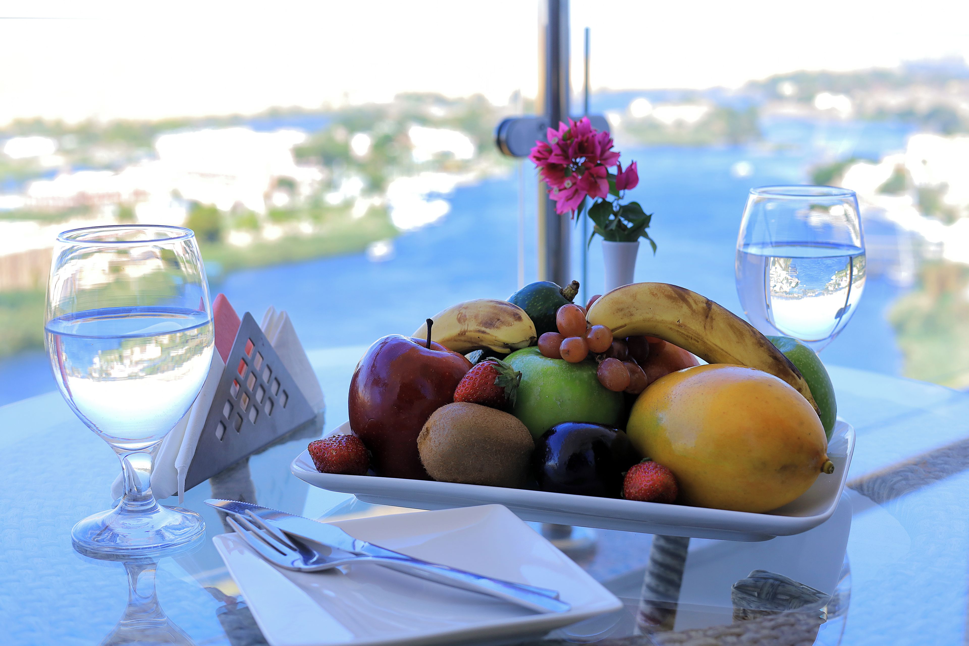 A table with a fruit bowl, a glass of water, and a flower.