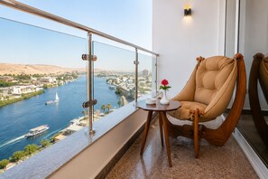 A balcony with a view of a river, a chair, a small table with a vase and a bowl, and a wall-mounted light fixture. - Blue Lotus Nile Suites (Aswan)