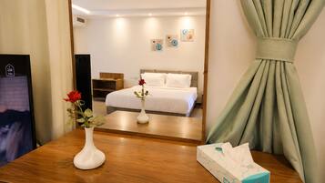 A hotel room with a bed, a mirror, a wooden table with a vase of flowers, a box of tissues, and artwork on the wall.