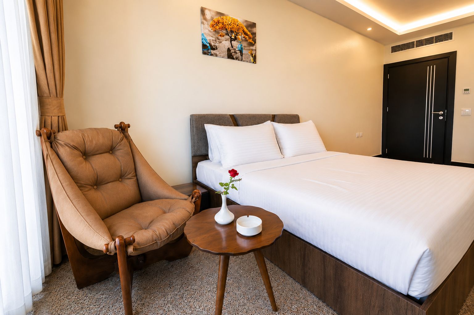 Habitación de hotel con una cama, una silla, una mesita con un florero y un cuadro en la pared.