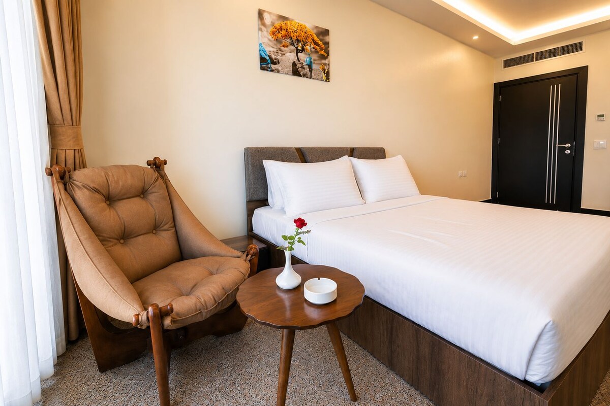 A hotel room with a bed, a chair, a small table with a vase, and a painting on the wall.