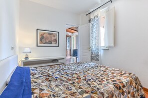 Un dormitorio con cama, mesita de noche con lámpara, cuadro enmarcado en la pared y cortina estampada.