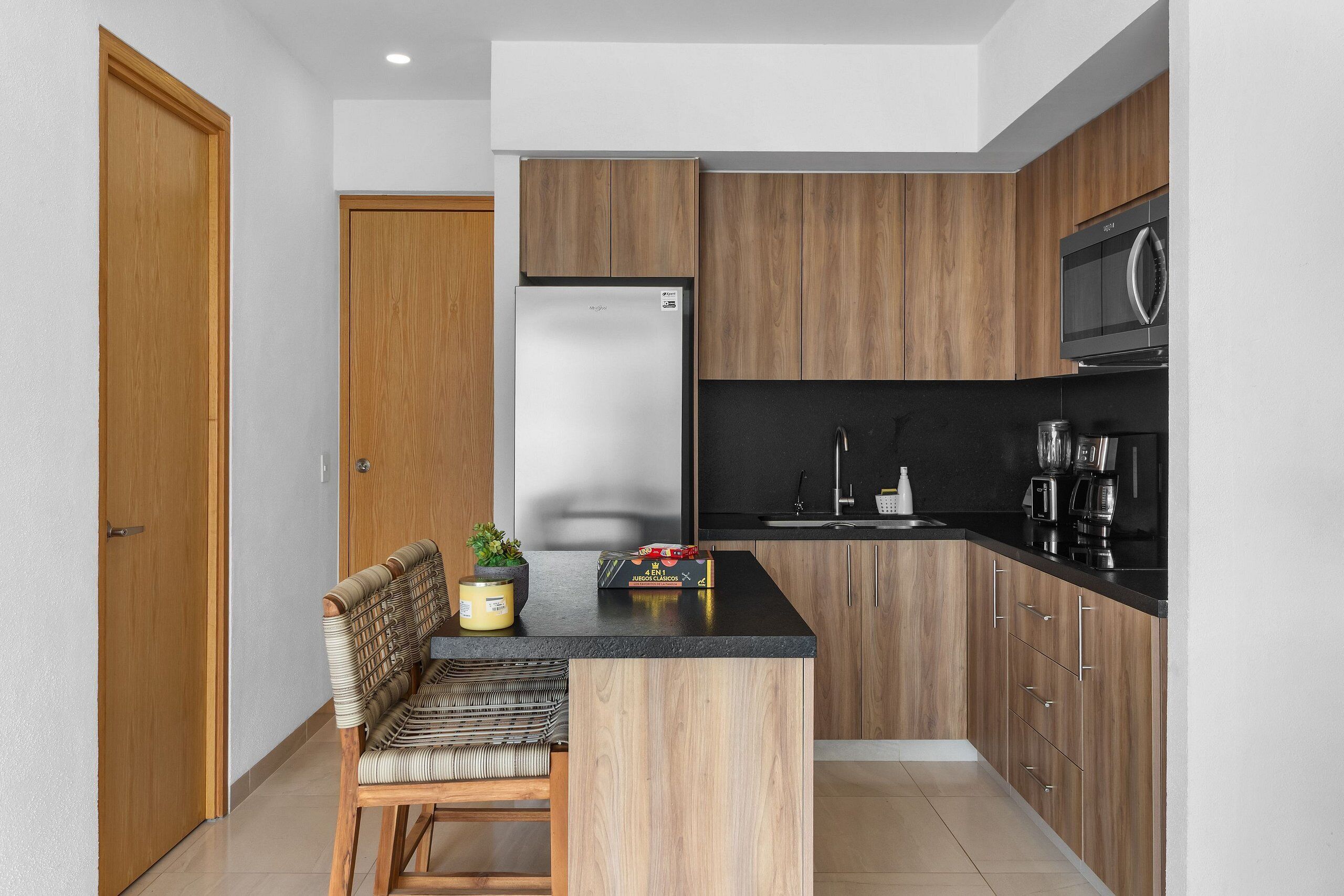 A compact kitchen with wooden cabinets, a black countertop, a built-in microwave, and a seating area with a chair and a small table.