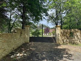 A gated entrance with stone walls and a metal gate, surrounded by trees and a clear sky.