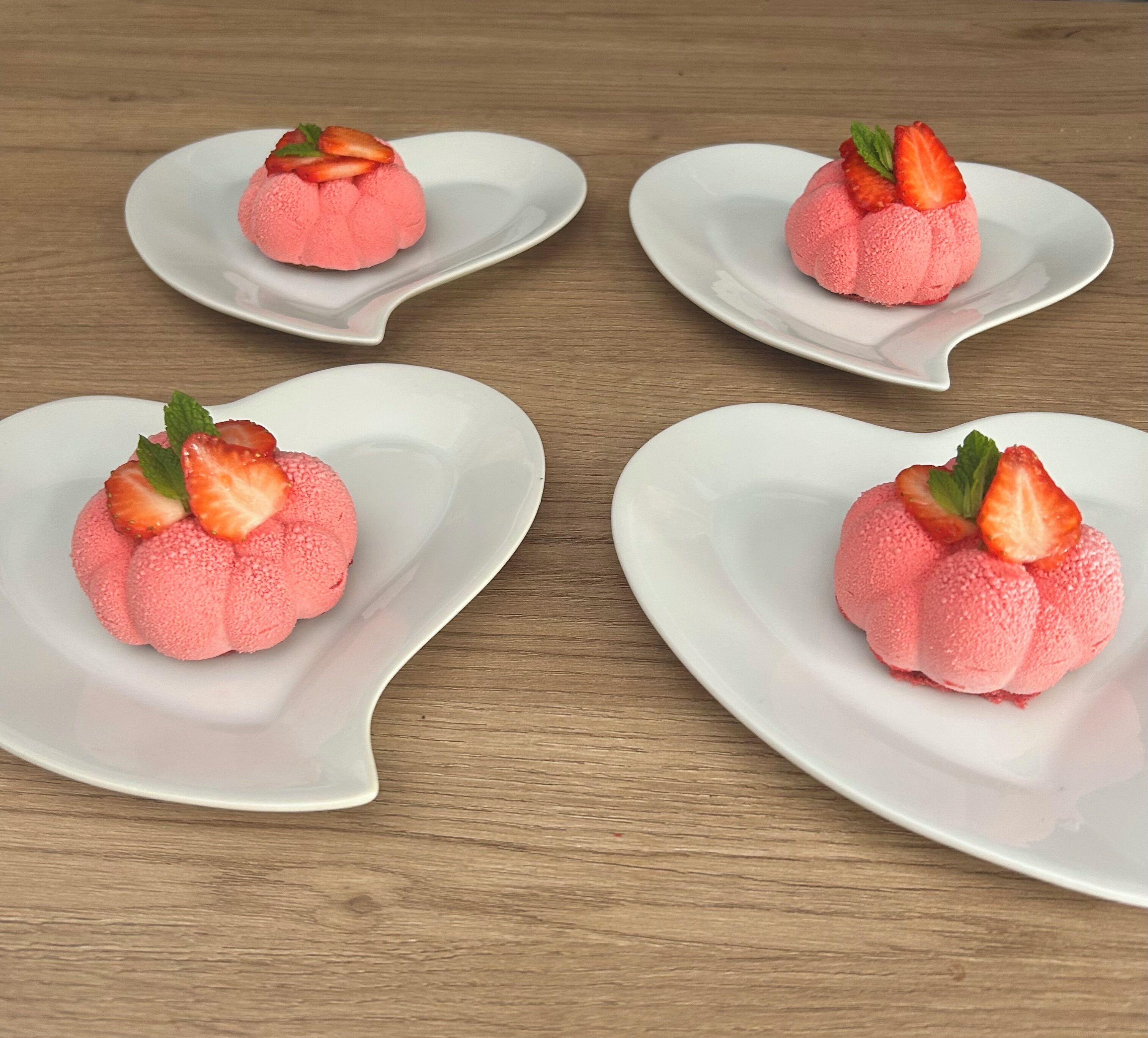 Four white plates with pink dessert shaped like strawberries on a wooden table.