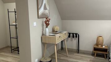 A modern loft bedroom with a sloped ceiling, a wooden desk with a vase and books, a woven basket, and a small side table with a lamp.