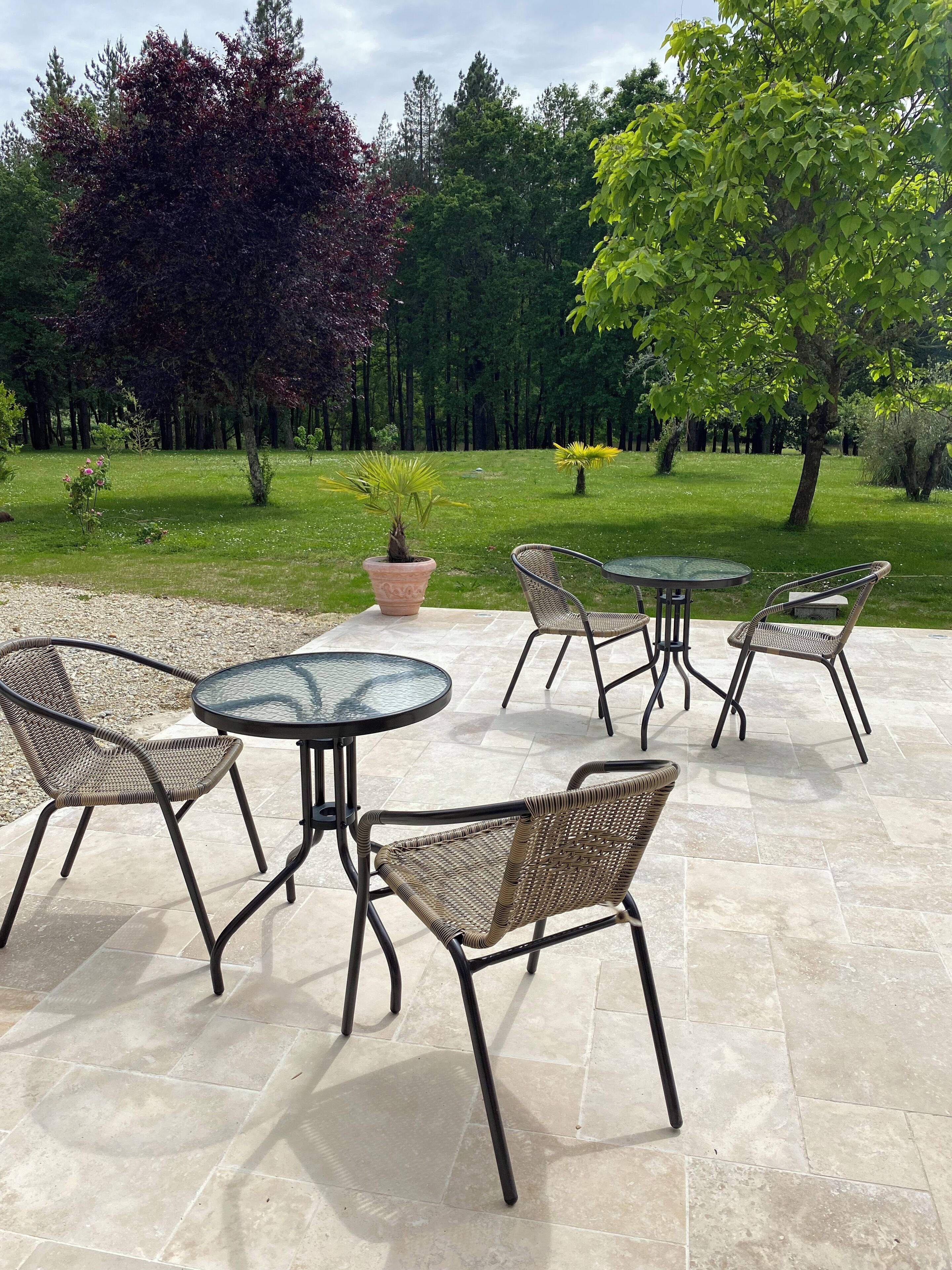Un patio al aire libre con mesas redondas y sillas de mimbre, rodeado de césped y árboles.
