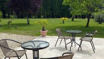 Un patio al aire libre con mesas redondas y sillas de mimbre, rodeado de césped y árboles.