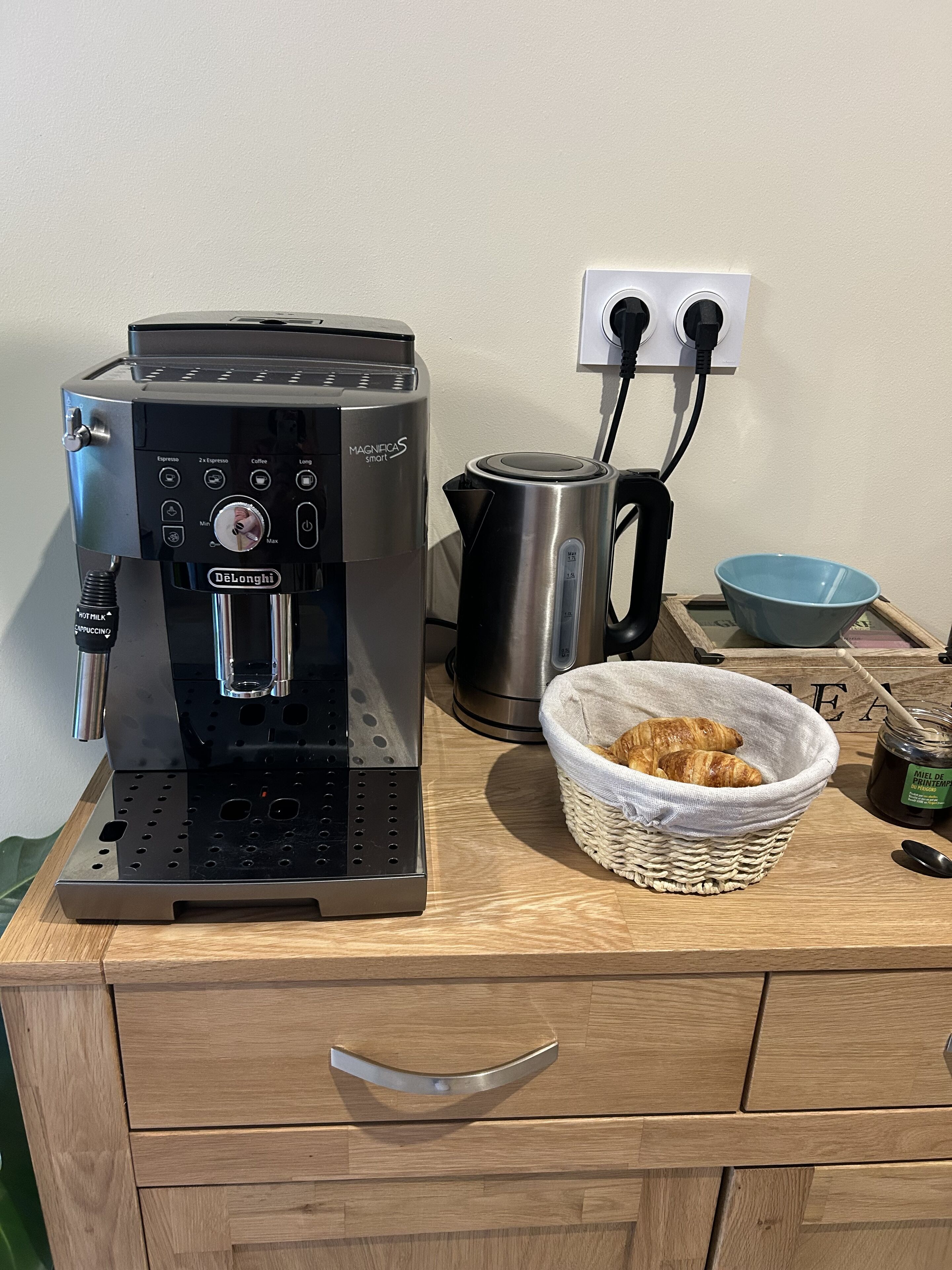 Kahvinkeitin, vedenkeitin, kori täynnä croissantteja ja purkki hilloa puisella työtasolla.