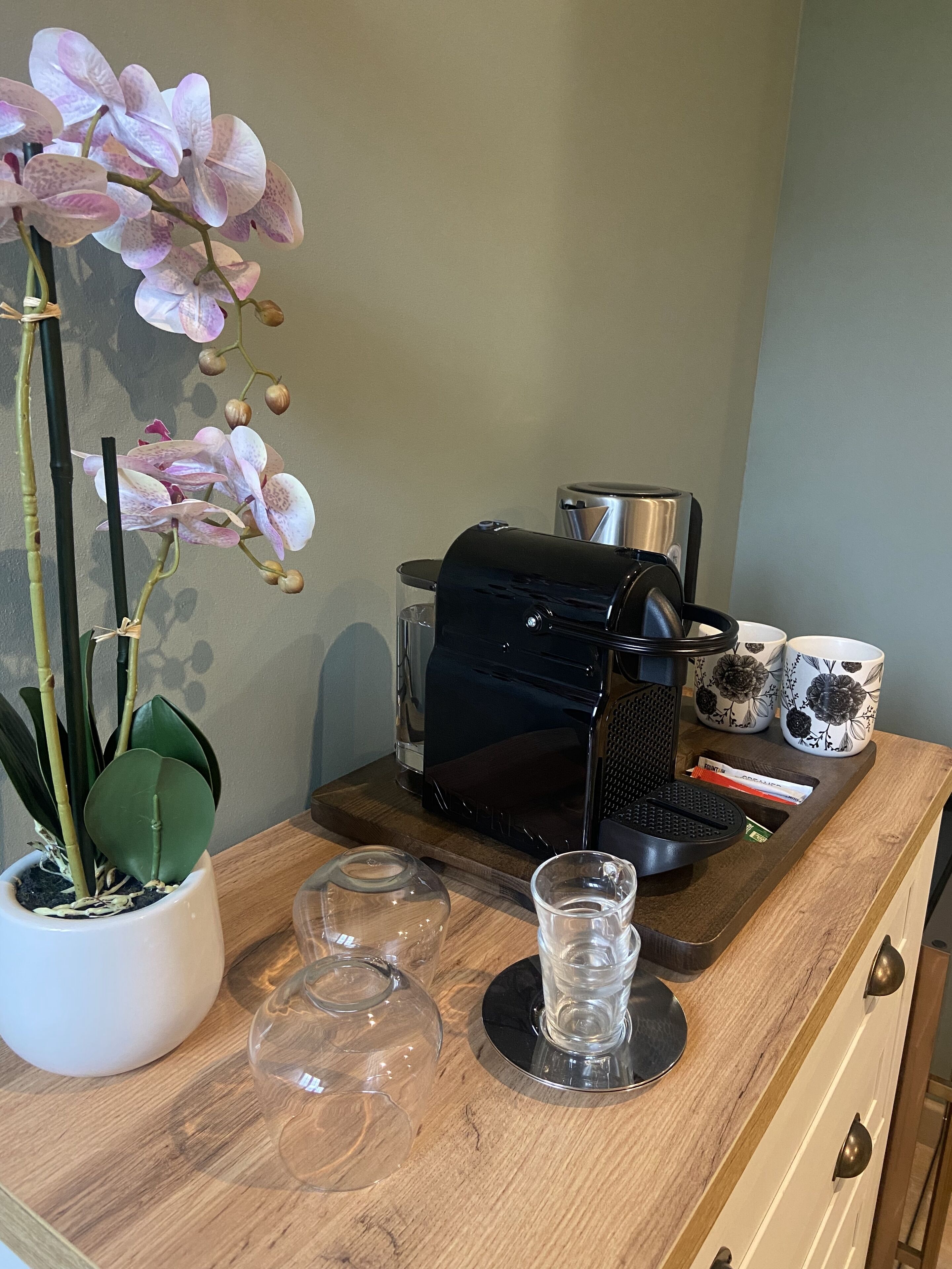 A coffee machine, two cups, a potted orchid, and a mug on a wooden counter.