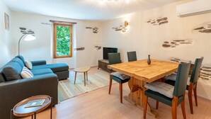 Een moderne woonkamer met een bank, een eettafel, stoelen, een televisie en een raam met uitzicht op bomen.