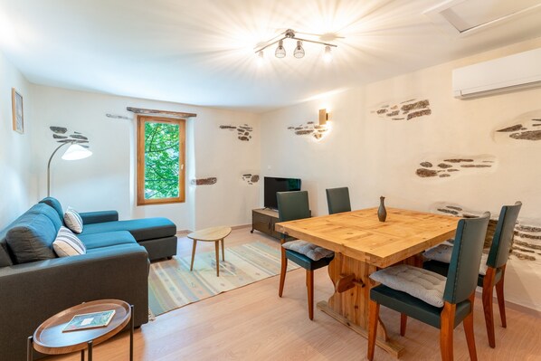 A modern living room with a sofa, a dining table, chairs, a TV, and a window with a view of trees.