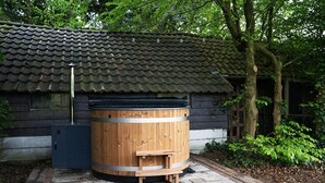 Un sauna traditionnel en bois, doté d’un toit en métal, situé dans un jardin avec une structure de toit en tuiles et un arbre à proximité.
