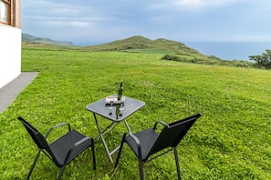 A grassy outdoor area with a table and two chairs, overlooking a hilly landscape and the sea.