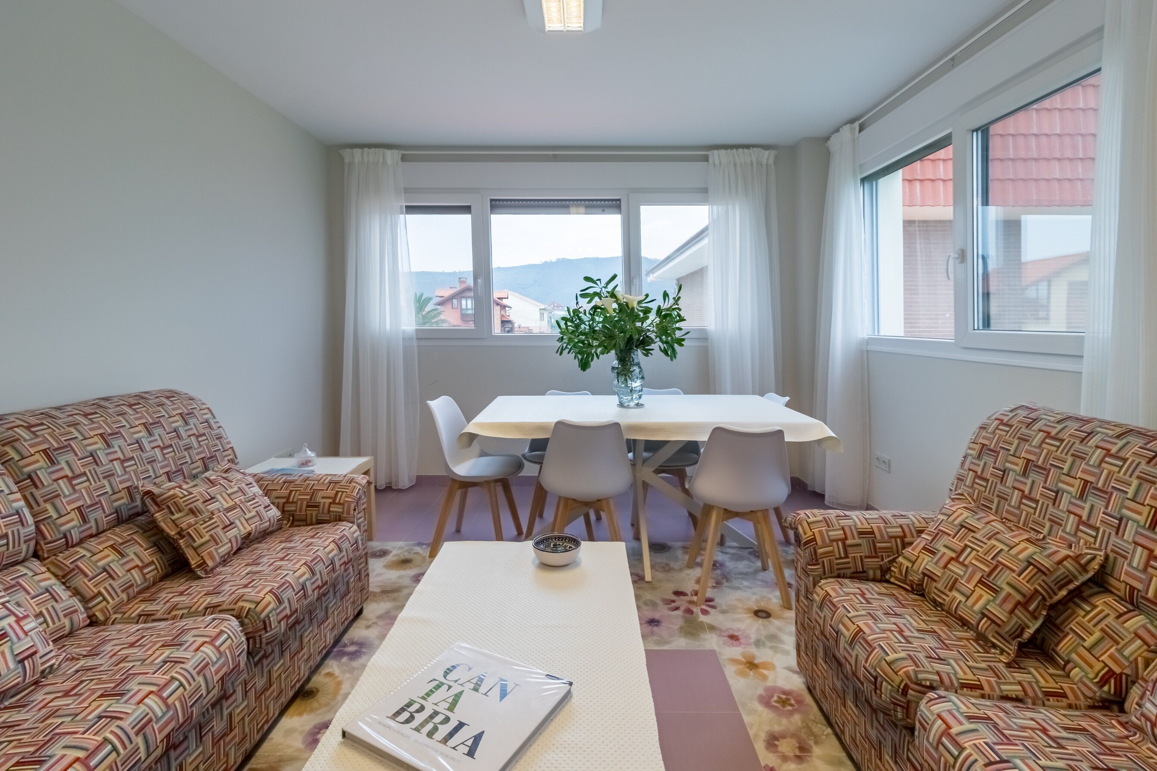 Un salon avec des canapés à motifs, une table à manger avec des chaises, un vase rempli de fleurs et un livre posé sur la table basse.