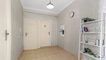 A hallway with a white door, a white shelf with books and a plant, and a clock on the wall.