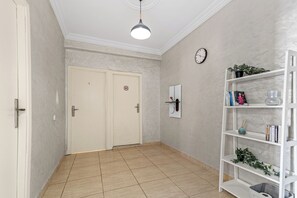 A hallway with a white door, a white shelf with books and a plant, and a clock on the wall. - Massira Social Living (Saâda)