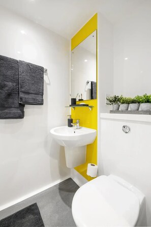A modern bathroom with a white sink, a yellow accent wall, a mirror, a toilet, and a black towel hanging on the wall. - Student Only Compass (Birmingham)