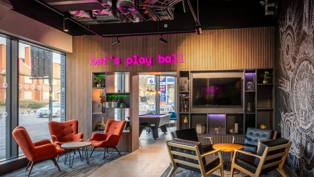 Ett modernt inomhusutrymme med ett biljardbord, orangefärgade stolar och en neonskylt med texten ”låt oss spela”.