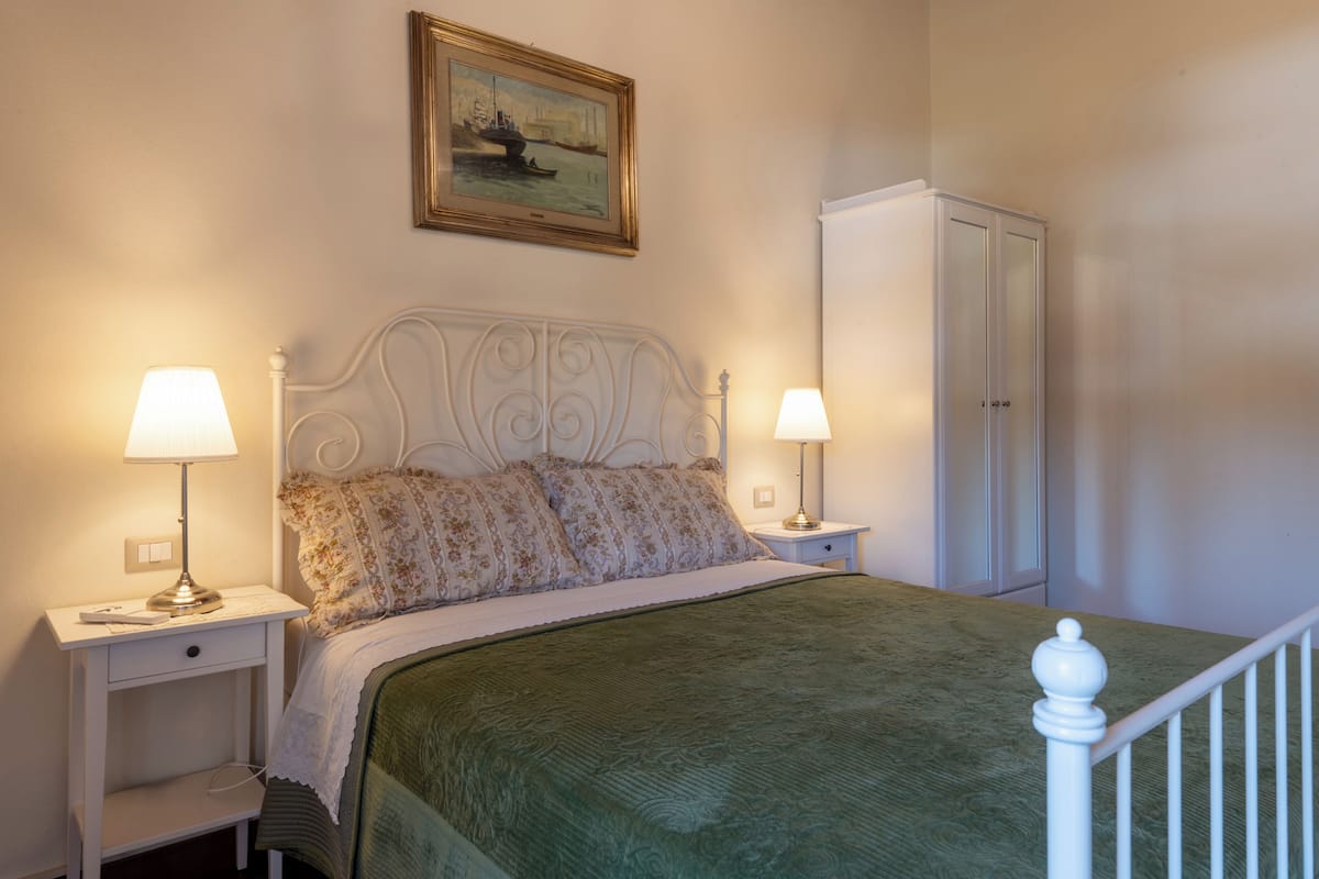 A neatly made bed with floral pillows, two bedside lamps, a framed picture on the wall, and a wardrobe.